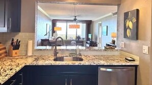 A kitchen with a granite countertop, stainless steel sink, and dark cabinets. There is a dining area visible through the kitchen, featuring a sofa and a dining table.