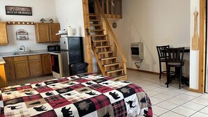A cozy bedroom with a bed, a wooden staircase, a kitchen area, and a dining table with chairs.