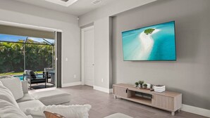 A modern living room with a large TV, a sofa, a coffee table, and a view of a pool and greenery outside. - Biohackers Retreat - Heated Pool, Spa, Sauna, Gym (Cape Coral)