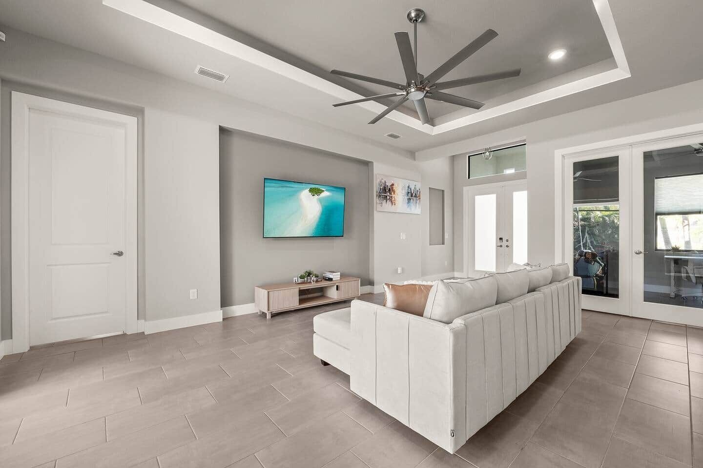 Un salón amplio con un sofá blanco modular, un ventilador de techo, un televisor de pantalla plana y un ventanal corredero que da a un área exterior.