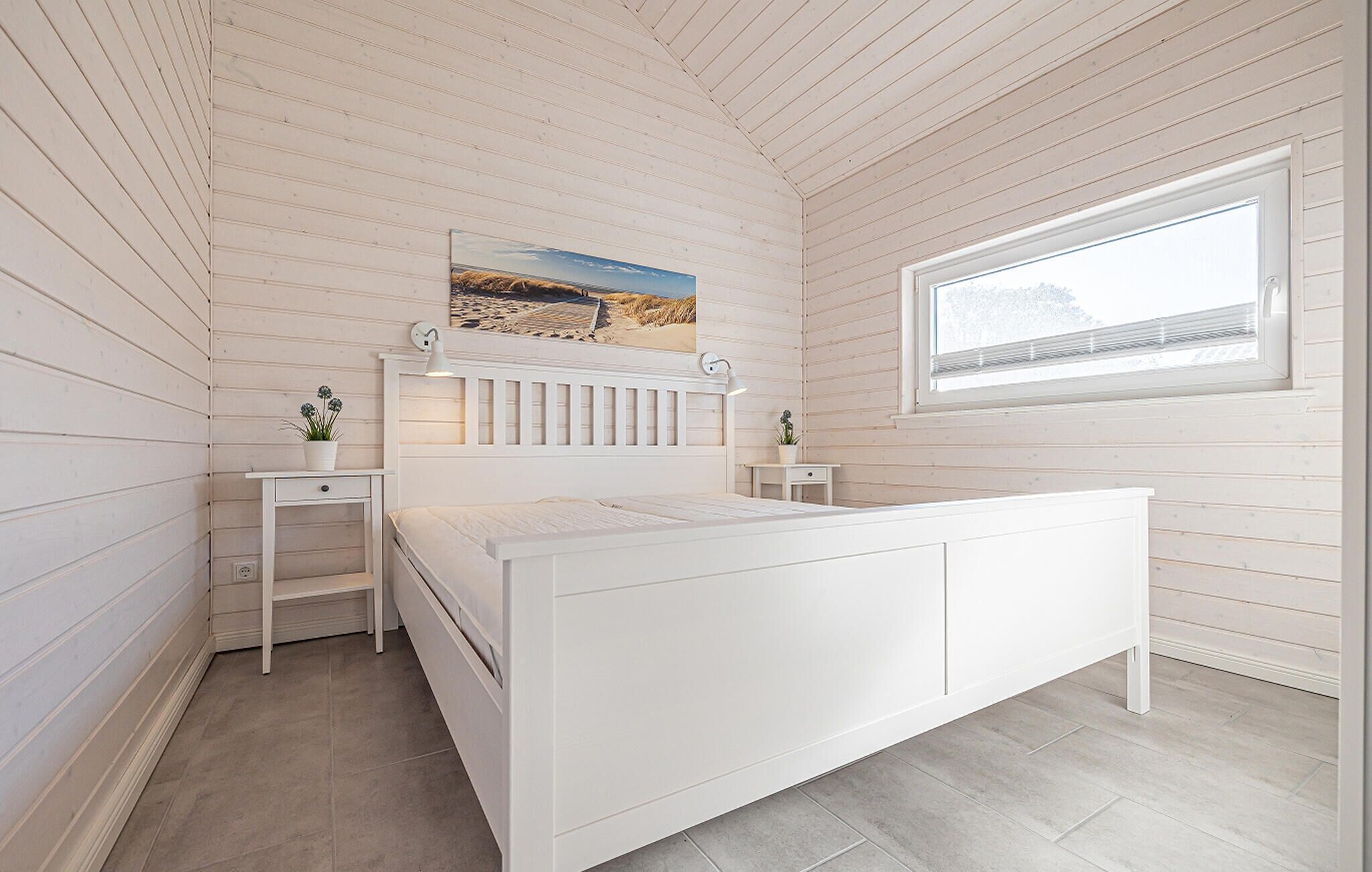 A bedroom with a white bed, a nightstand, a small table, a window, and a beach painting on the wall.