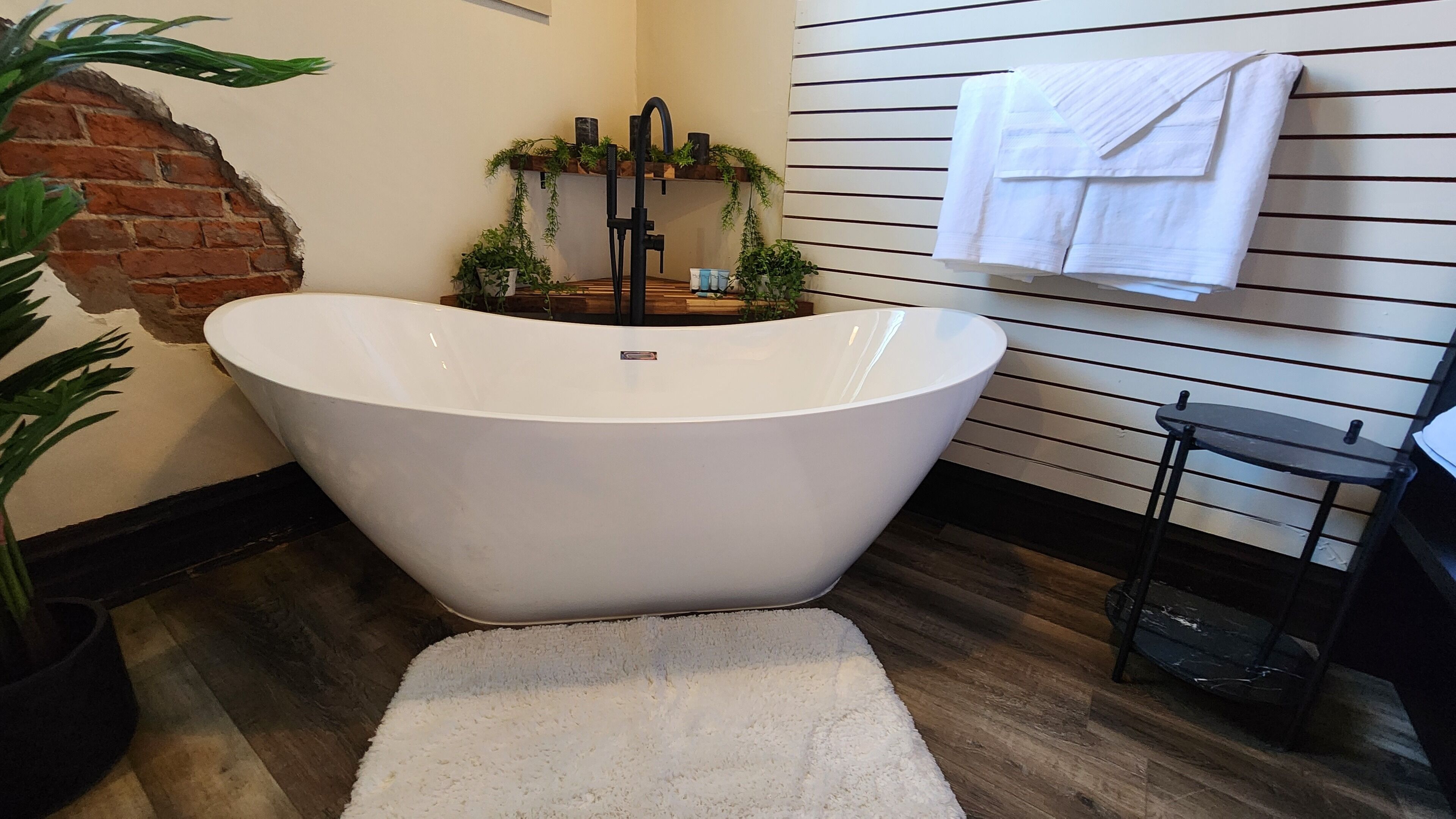 A modern bathroom with a freestanding bathtub, a brick accent wall, and a wooden floor.