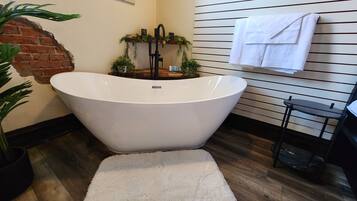 A modern bathroom with a freestanding bathtub, a brick accent wall, and a wooden floor.
