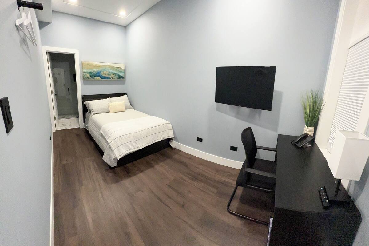 Ein Schlafzimmer mit Bett, Schreibtisch, Stuhl, Fernseher und einer Pflanze.