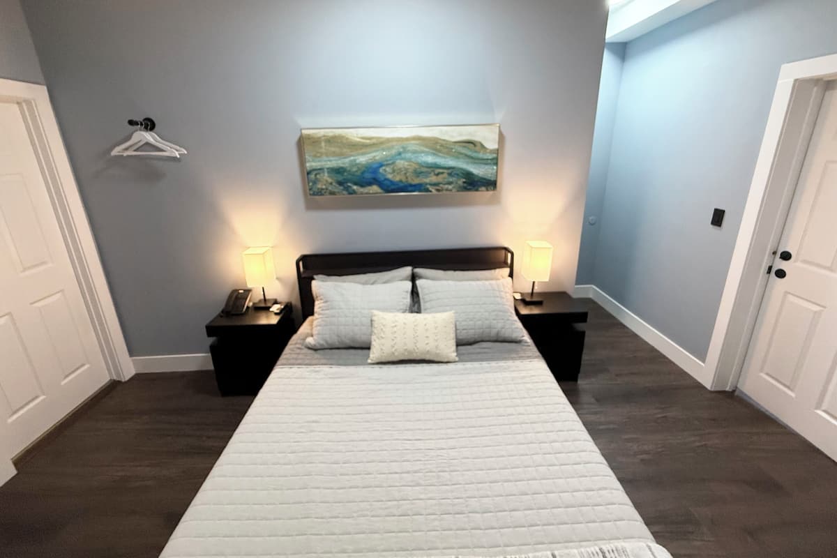 Ein Schlafzimmer mit einem großen Bett, zwei Nachttischen mit Lampen, einem Bild an der Wand und einer Tür links.