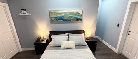 A bedroom with a large bed, two bedside tables with lamps, a painting on the wall, and a door on the left.