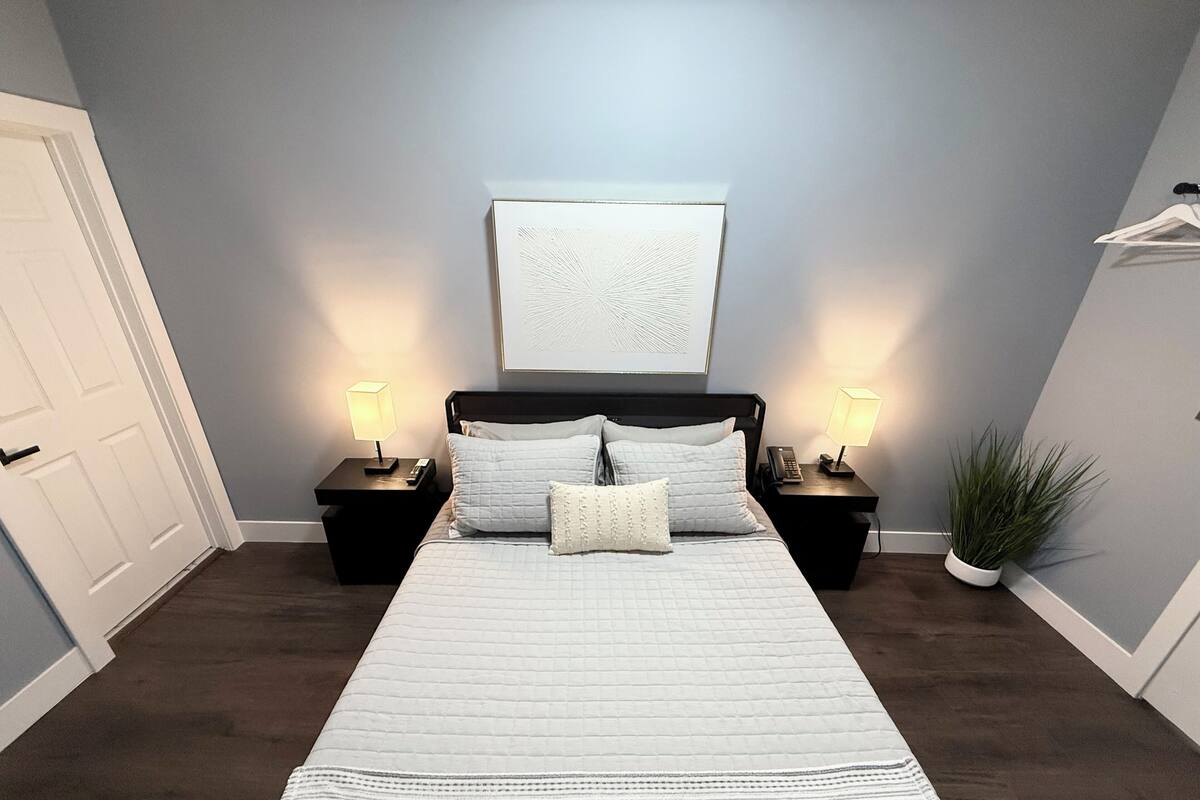Ein Schlafzimmer mit einem großen Bett, zwei Nachttischen mit Lampen, einer Tür, einem an der Wand hängenden Kunstwerk und einem Blumentopf.