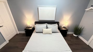 A bedroom with a large bed, two bedside tables with lamps, a door, a wall-mounted artwork, and a potted plant.