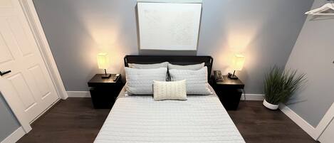 A bedroom with a large bed, two bedside tables with lamps, a door, a wall-mounted artwork, and a potted plant.
