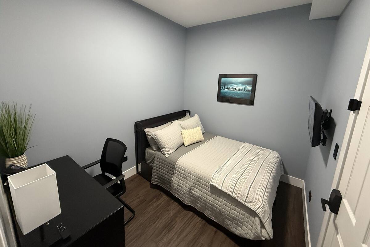 Ein Schlafzimmer mit einem Bett, einem Schreibtisch, einem Stuhl, einer Zimmerpflanze und einem gerahmten Bild an der Wand.