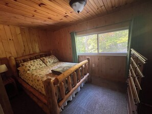 A wooden cabin bedroom with a bed, dresser, and window with a view of trees.