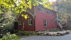 Une maison en bois rouge, avec une allée en gravier, entourée de rochers et d’arbres.