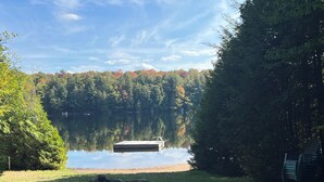 Un cadre paisible au bord d’un lac, avec une jetée s’avançant dans des eaux calmes, entouré d’arbres denses et d’une pile de bois de chauffage sur la rive herbeuse.