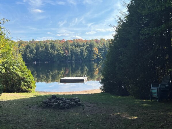 A serene lakeside setting with a dock extending into calm waters, surrounded by dense trees and a pile of firewood on the grassy shore.