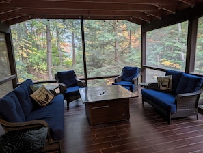 A covered outdoor seating area with wicker furniture and blue cushions, overlooking a forest. - Puchers Place - Spacious, waterfront home on a private non-motorized lake (Thendara)