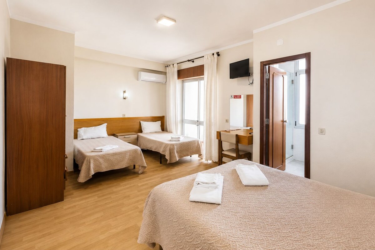 A hotel room with two single beds, a larger bed, a desk, and a TV.