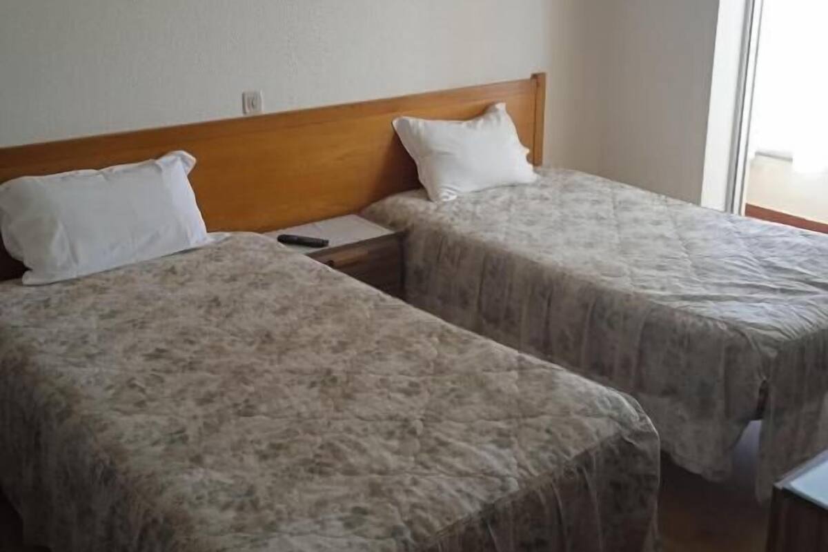 A room with two beds, each with a wooden headboard and white pillows. There is a nightstand between the beds, and a window to the right.