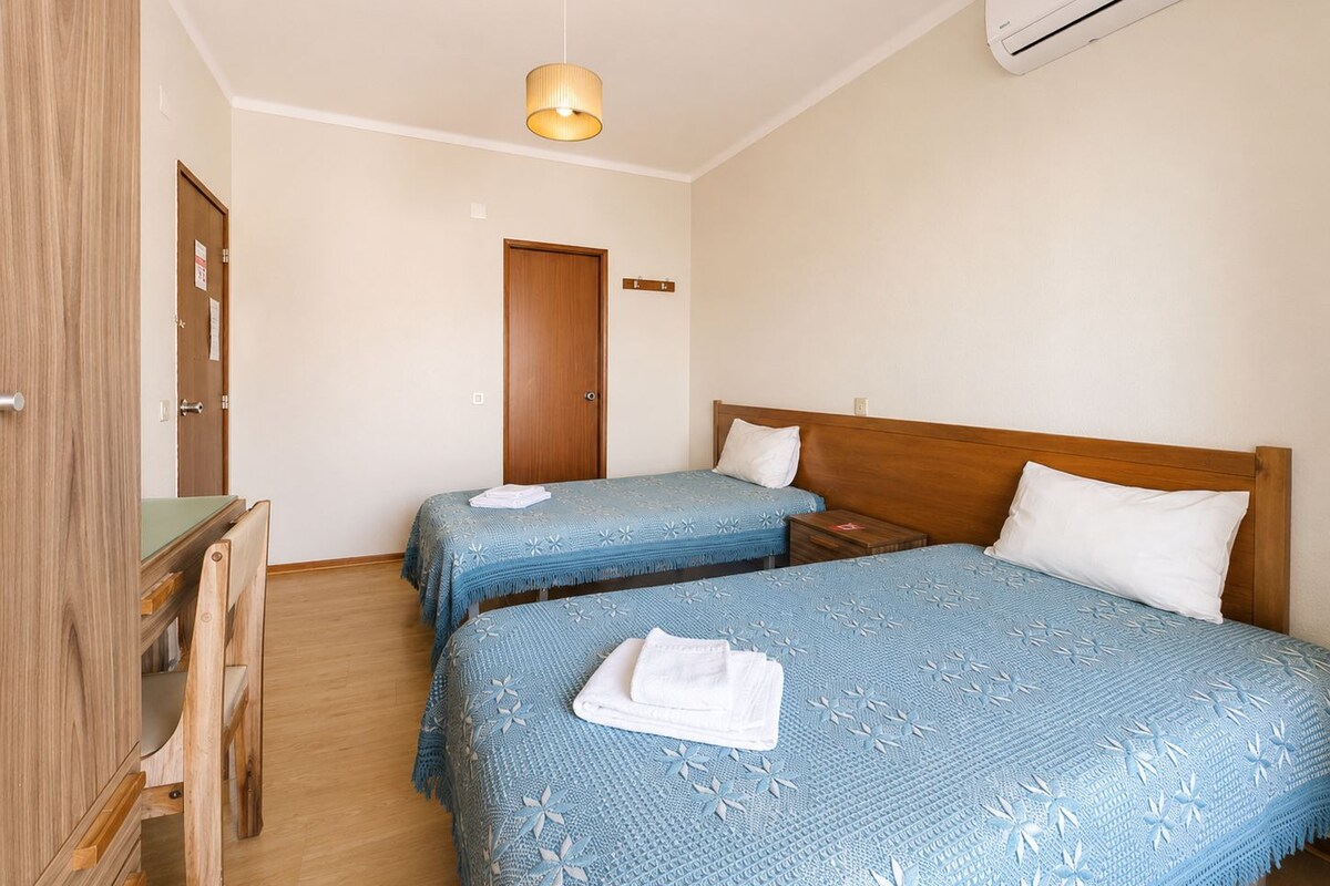 A hotel room with two beds, a wooden desk, a chair, a hanging lamp, and an air conditioning unit.