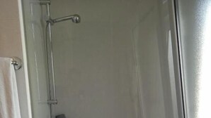 Une cabine de douche fermée par des parois en verre, avec une serviette blanche accrochée au mur, une petite étagère et un drain.