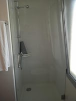 Un box de ducha con paredes de vidrio, una toalla blanca colgada en la pared, un estante pequeño y un desagüe.