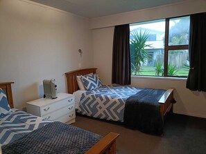A bedroom with two beds, a bedside table with a lamp, a dresser, and a window with a view of a backyard. - Ōhope Beach 3 Bedroom Holiday House (Ōhope)