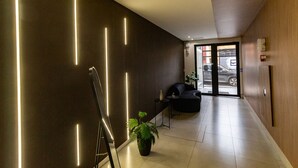 A modern hotel lobby with a dark wall, a potted plant, a black sofa, and a glass door leading outside.