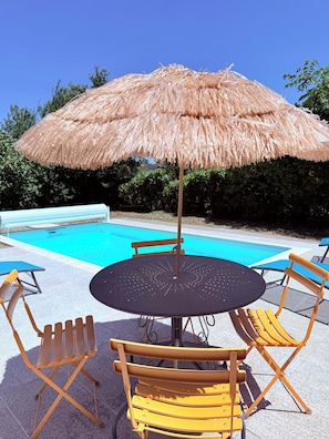A poolside setting with a round table, four yellow chairs, and a thatched umbrella.
