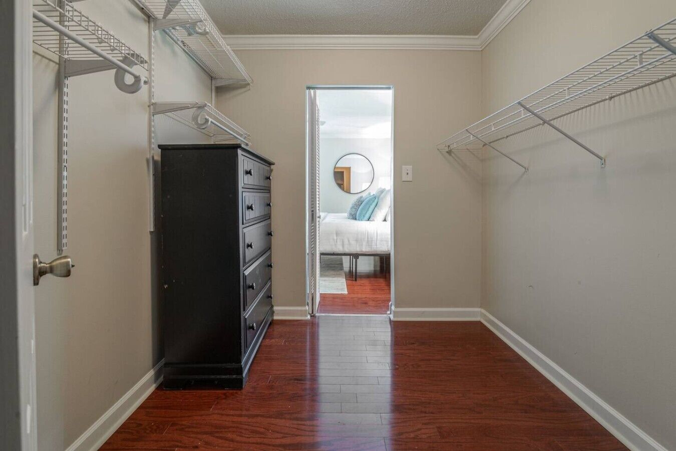 Un couloir étroit au plancher de bois, une commode noire et un lit avec un grand miroir rond et des coussins dans la pièce adjacente.