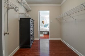 A narrow hallway with wooden flooring, a black dresser, and a bed with a round mirror and pillows in the adjacent room. - Spacious Retreat Minutes from Samford (Birmingham)