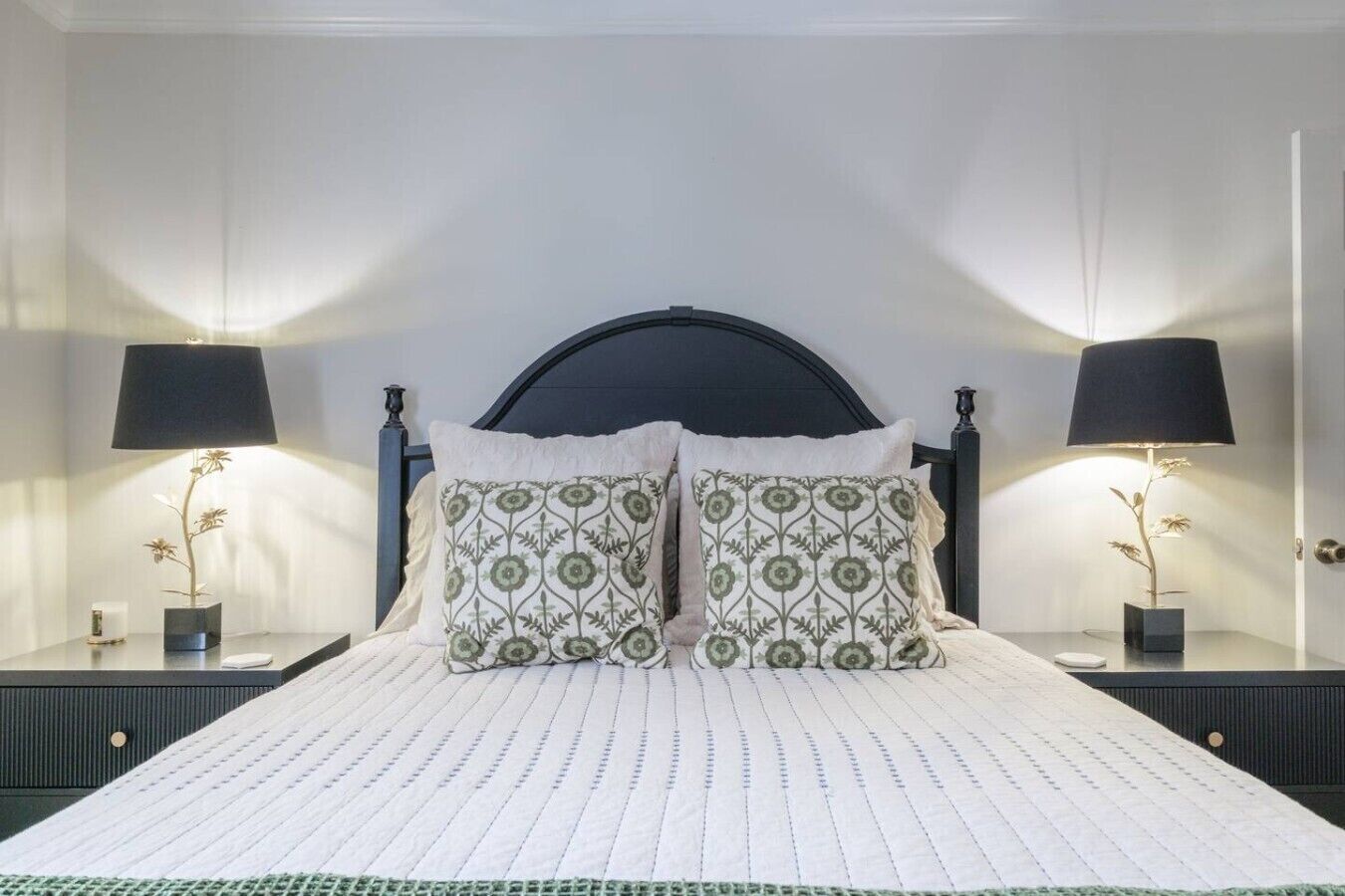 Un lit bien fait, avec une couette blanche et deux oreillers, de part et d’autre duquel se trouvent deux tables de chevet ornées de lampes et de vases décoratifs.