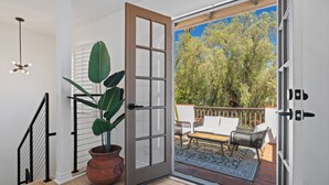 An open sliding glass door leading to an outdoor patio with a wooden deck, a white sofa, and a dining table. - Luxury 5BR Villa - Pool, Spa, Movie Theater, Game Room, Bar, & Outdoor Dining (La Mesa)