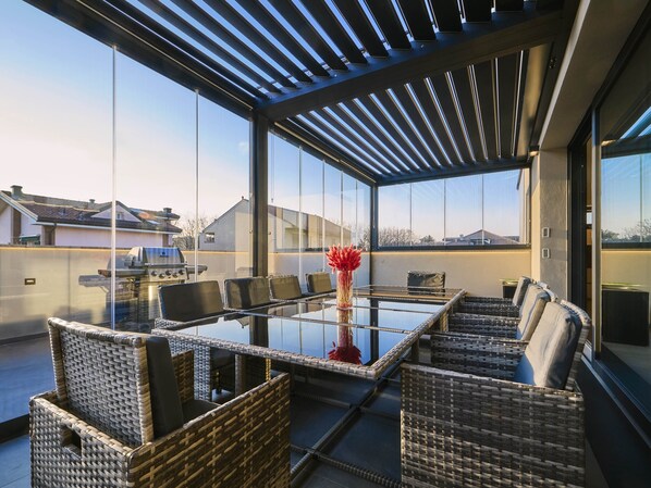 Een modern buitenontbijtgedeelte met een glazen tafel en rieten stoelen, met uitzicht op een woonwijk.