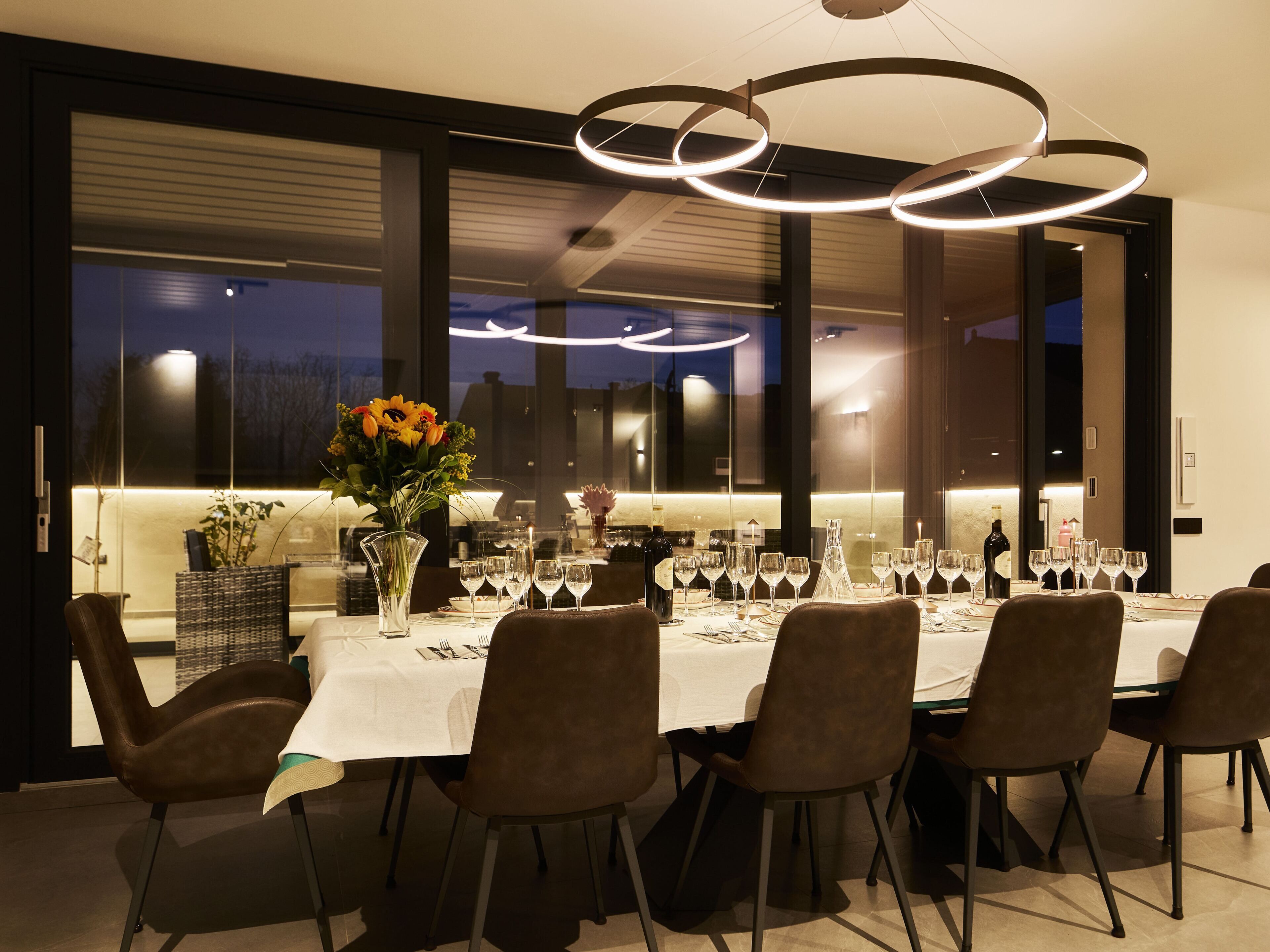 A modern dining area with a long table set for a meal, large windows, and contemporary lighting fixtures.