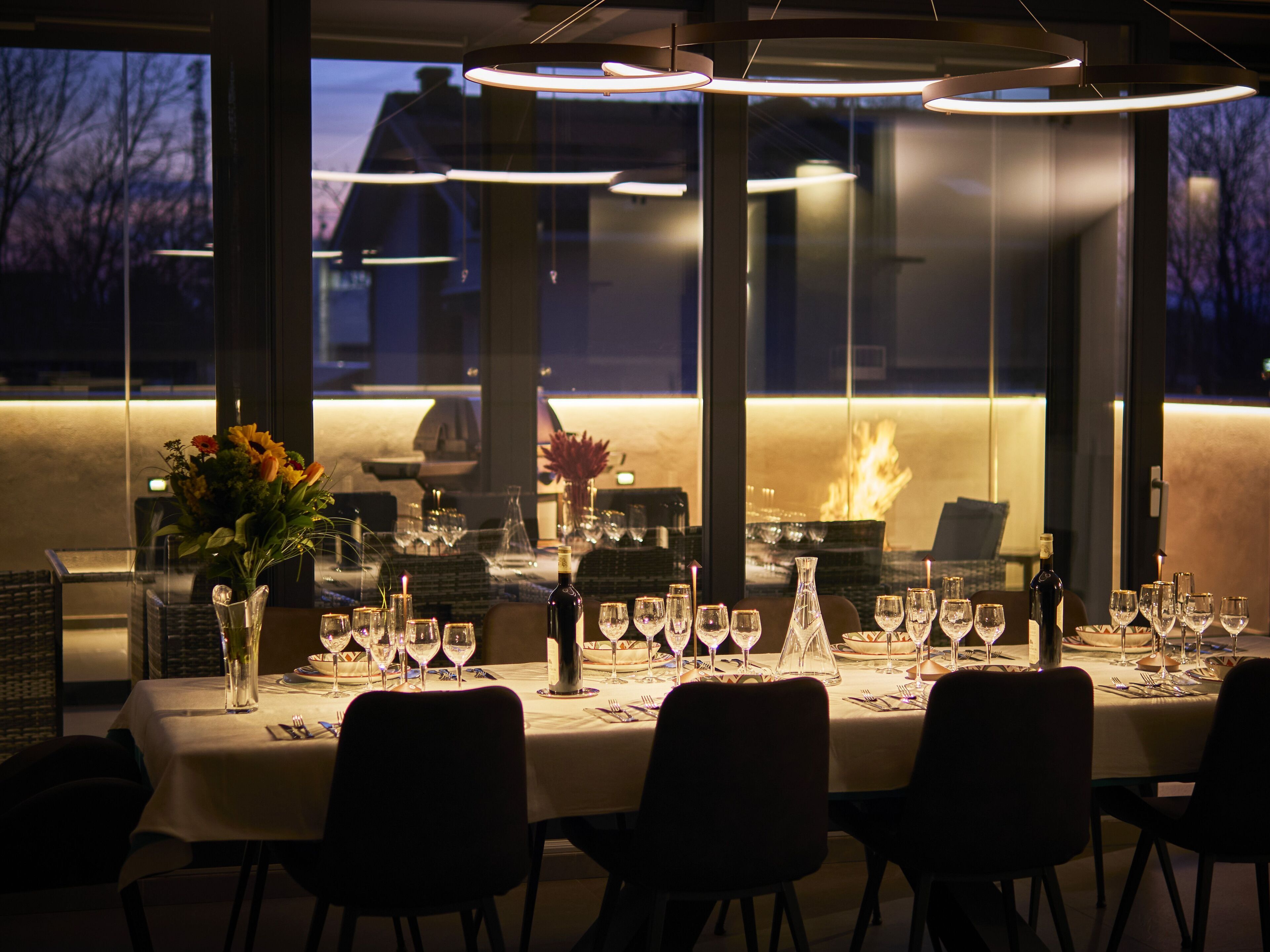 Un restaurant avec une longue table dressée pour un repas, de grandes fenêtres et des luminaires modernes.