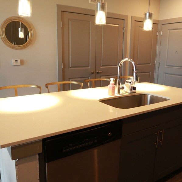Una cocina moderna con encimera blanca, pileta de acero inoxidable y lavavajillas. Hay sillas de madera y un espejo redondo en la pared.