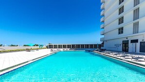 A rectangular swimming pool with clear blue water, surrounded by lounge chairs and a building with multiple floors and windows. - Fully Remodeled 2 Bedroom with Direct Beach Access (Miami Beach)