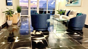 A modern living room with a glass coffee table, two blue armchairs, a white sofa, and a large window offering a city view. - Fully Remodeled 2 Bedroom with Direct Beach Access (Miami Beach)