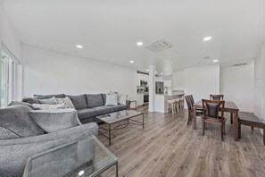 Ein modernes Wohnzimmer mit einem grauen Sofa, einem Glastisch und einem Essbereich mit Holzsesseln und einem Tisch.