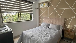 Un dormitorio con cama, mesita de noche, aire acondicionado instalado en la pared y ventana con persianas.
