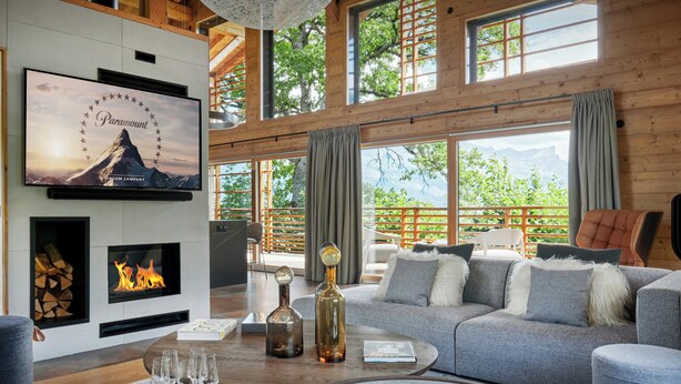 A spacious living room with a fireplace, a large TV mounted on the wall, and a view of the outdoors through large windows. - Chalet RevAlp - All-season holidays with private sauna - OVO Network (Combloux)