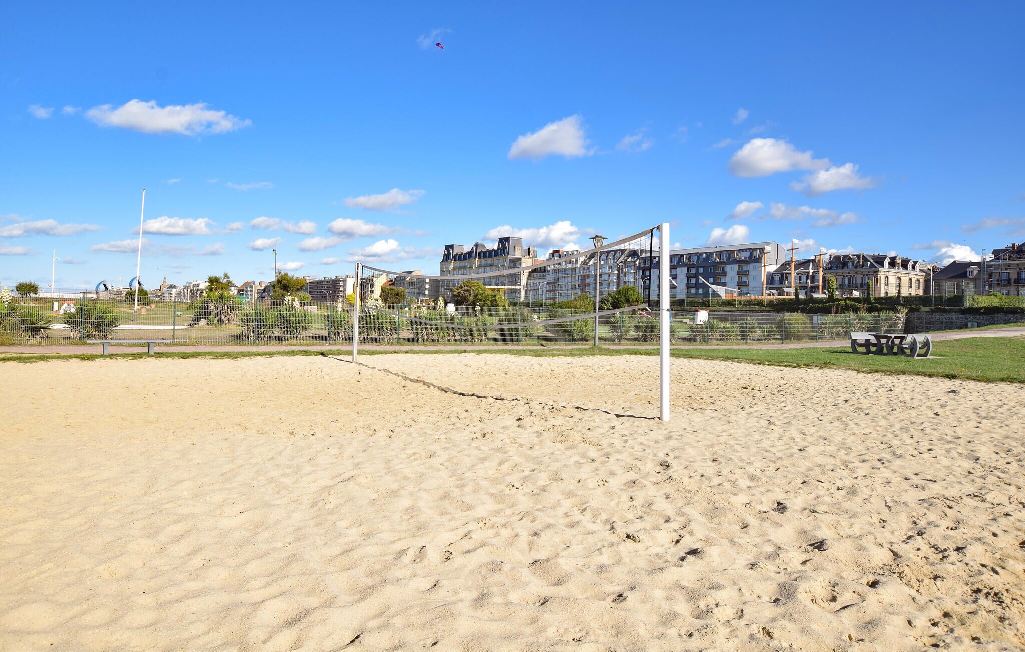 Ein Beachvolleyballfeld aus Sand mit Netz, umgeben von einer Umzäunung, mit Gebäuden im Hintergrund.