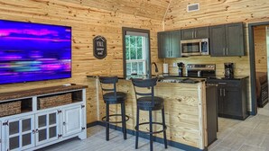 A cozy cabin interior with a wooden ceiling, a flat-screen TV, a kitchen area, and a bar stool.
