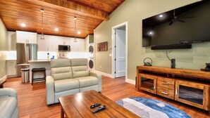 A spacious living area with a wooden ceiling, a large flat-screen TV, a sofa, a coffee table, and a kitchen in the background. - Rushing Waters Cabin (Sylva)