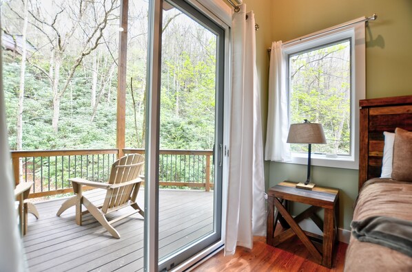 A room with a wooden deck, a chair, a bedside table with a lamp, and a bed. - Rushing Waters Cabin (Sylva)