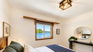 Um quarto com uma cama, uma janela com vista para um lago, uma mesa de cabeceira com abajur e um espelho na parede.