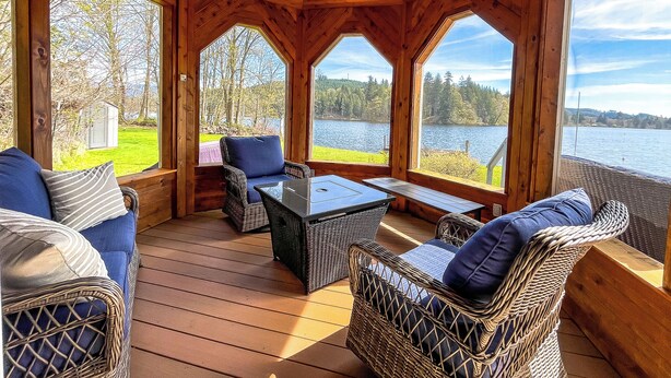 Um deck de madeira com móveis de vime, uma mesa de vidro e janelas amplas com vista para o lago.