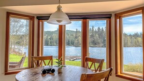 Uma área de refeições com uma mesa redonda, quatro cadeiras, um lustre e grandes janelas com vista para um lago.