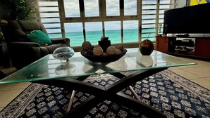 A living room with a glass coffee table, a bowl of fruit, and a view of the ocean through large windows.