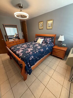 Quarto com cama de madeira, mesa de cabeceira com abajur, cômoda e espelho.