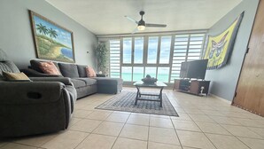 A living room with a sofa, a coffee table, a TV, and a view of the ocean through large windows.